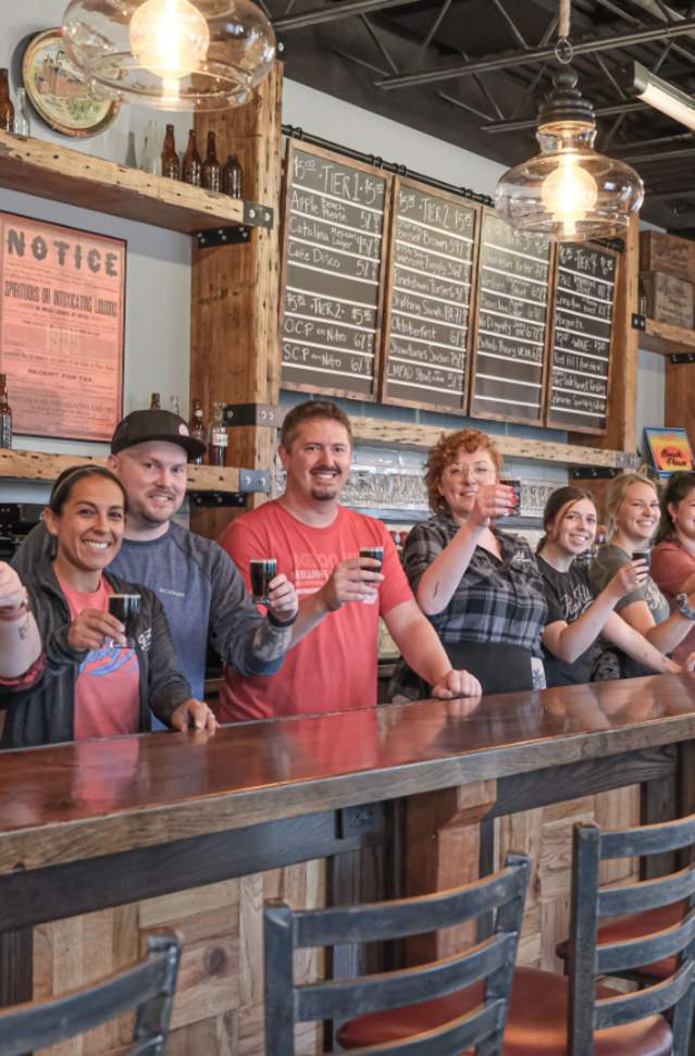 A group of people at a brewery bar raise small beer glasses in a toast. They stand by a long wooden counter with menus on the wall, looking cheerful.