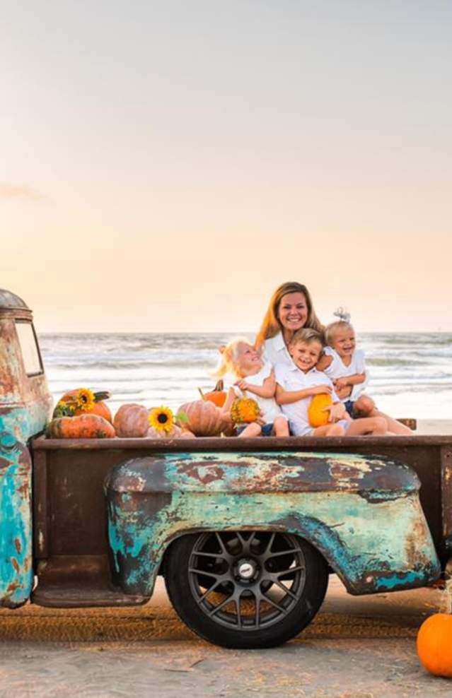 An old truck sits on the beach surrounded by pumpkins. A family sits in the bed of the truck and a logo overlaid on the photo reads "Beachtober."