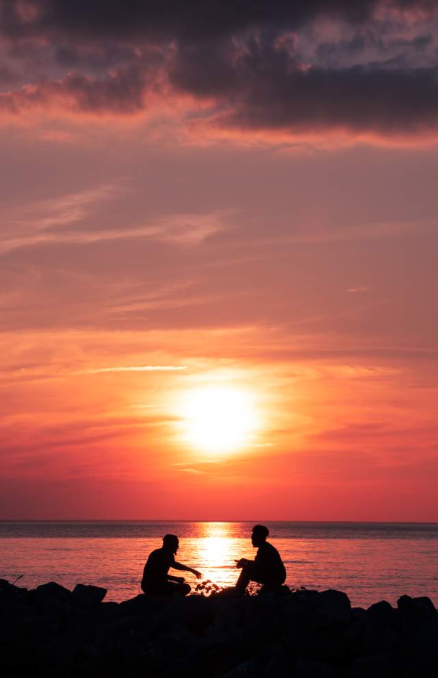 2 people sit on rocks on the shore of the James River silhouetted by a sunset