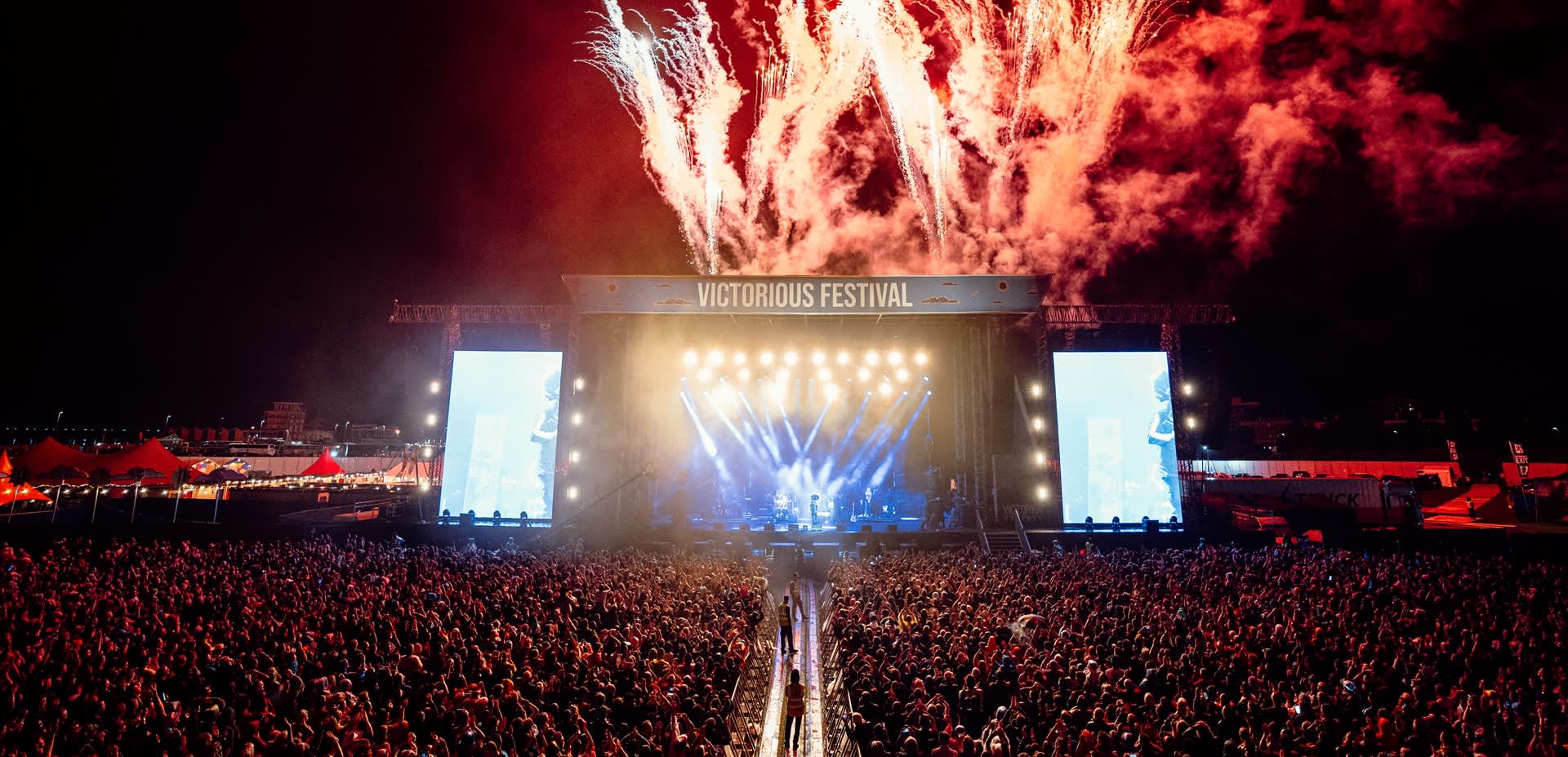 Photograph showing the culmination of a headline set at Victorious Festival, with red fireworks going off behind the stage and thousands of people gathered in front