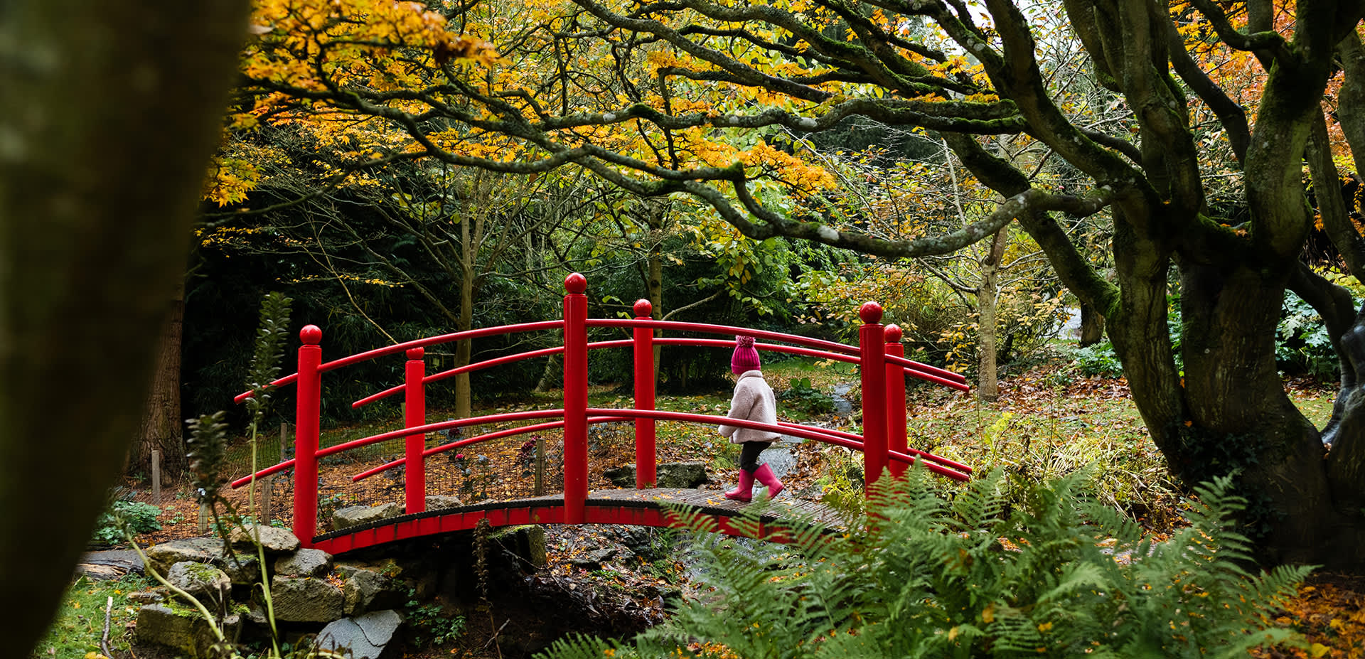 Child in bobble hat and pink coat crossing a bridge at Batsford Arboretum