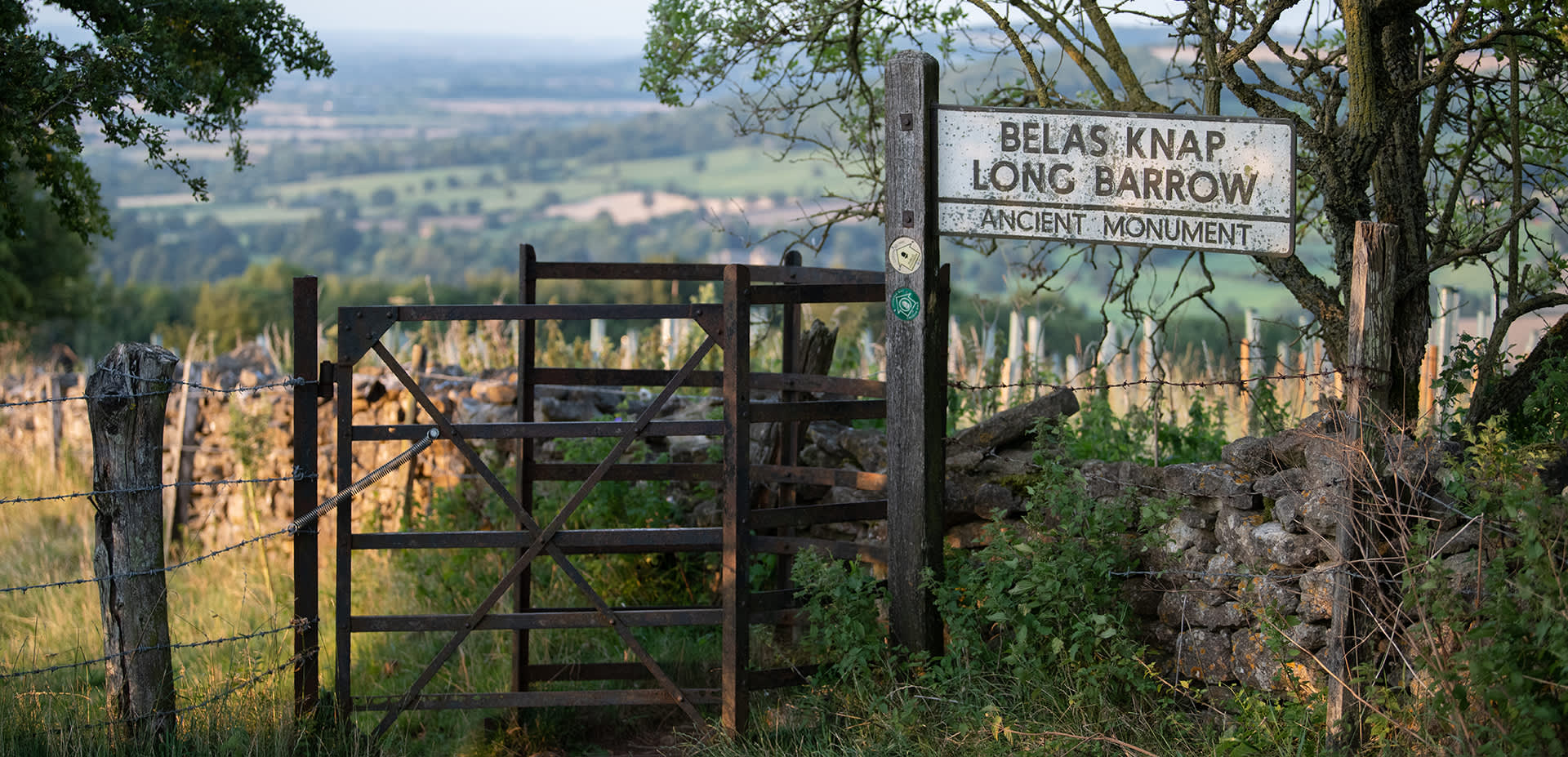 Kissing gate with footpath sign to Belas Knap with a stone wall ahead and behind a view of the valley