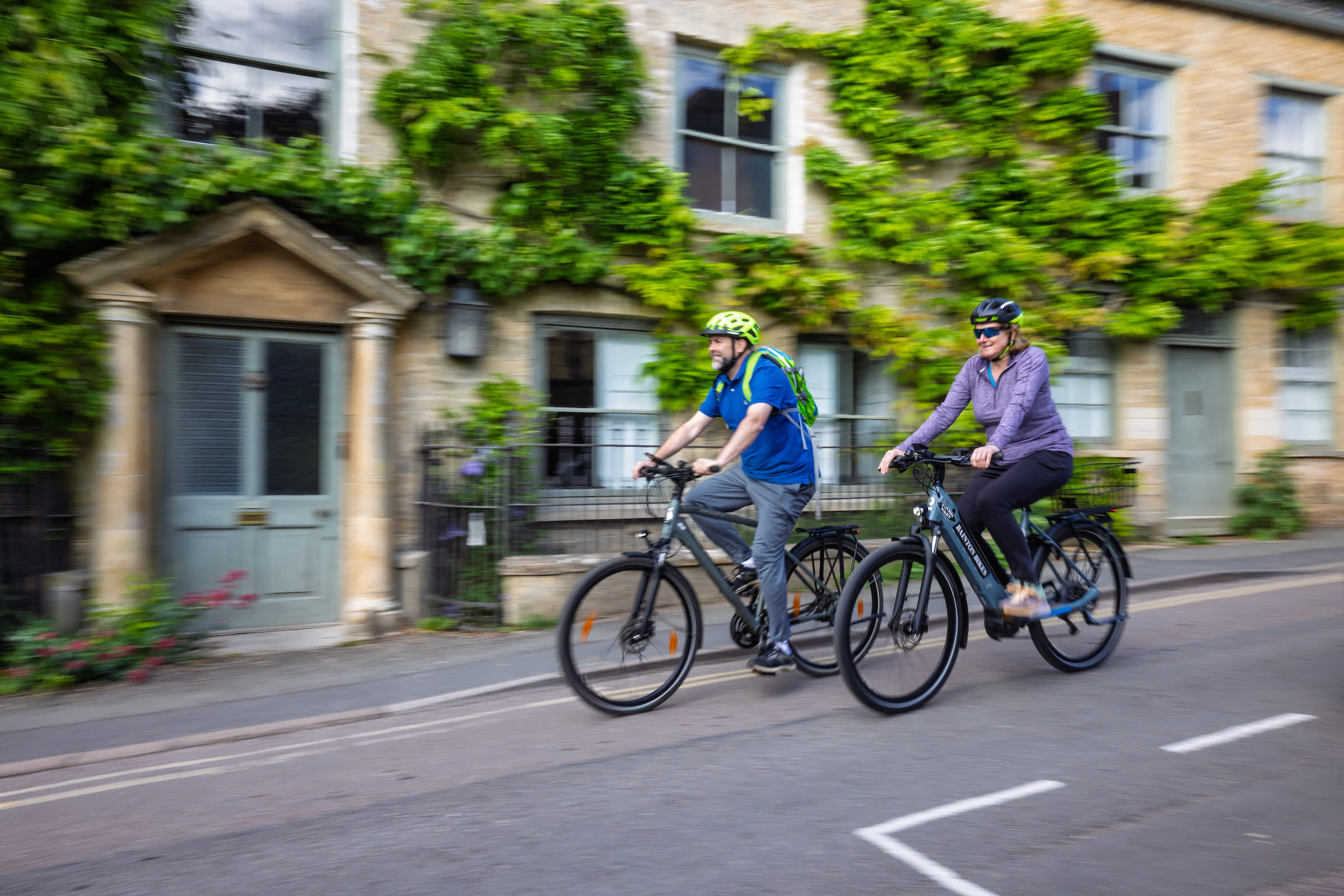 Two cyclists riding bikes through picturesque Charlbury, passing stone cottages covered with greenery