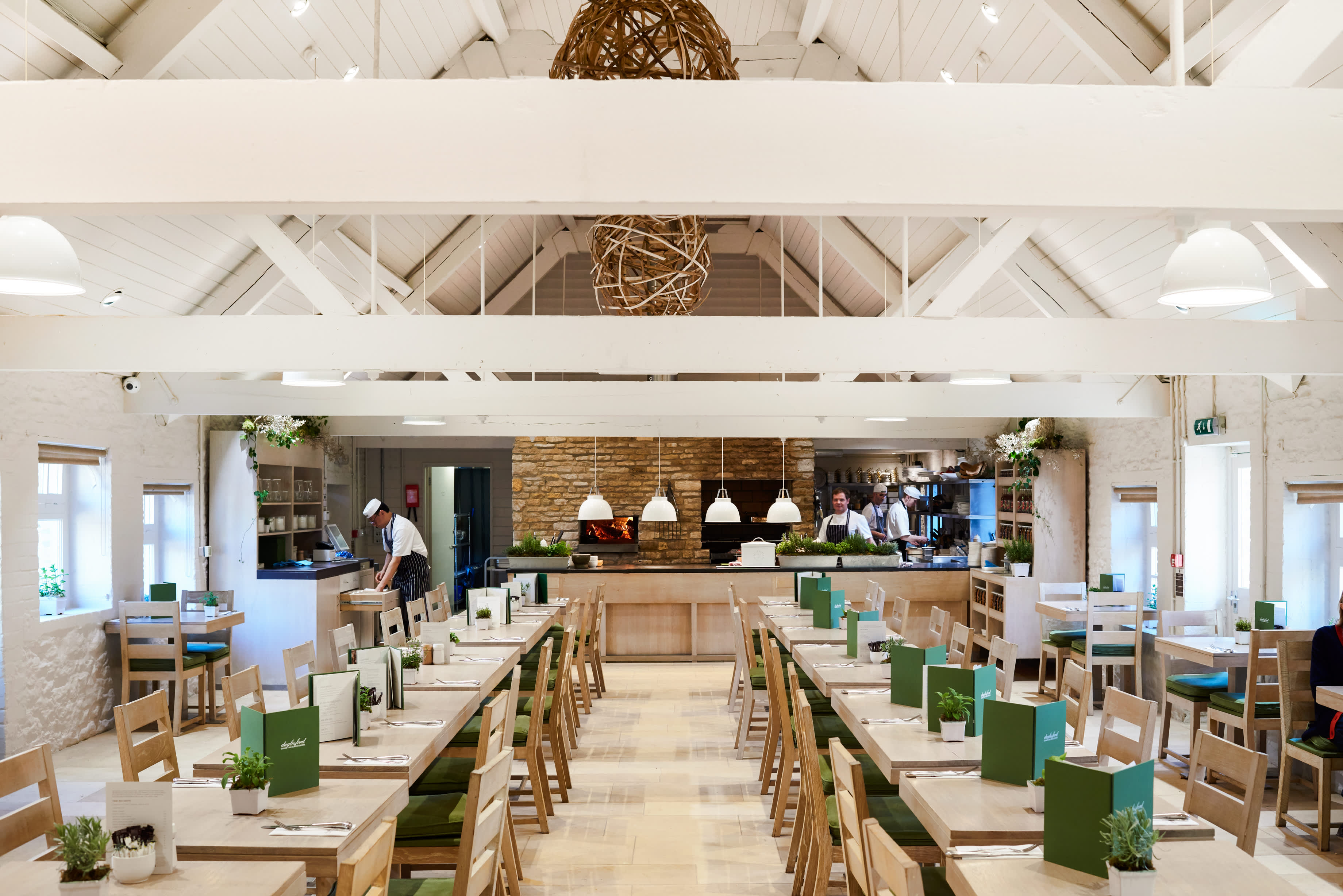 Rows of light wooden dining tables with green menus lead to an open kitchen where chefs are preparing for dinner at Daylesford in the Cotswolds