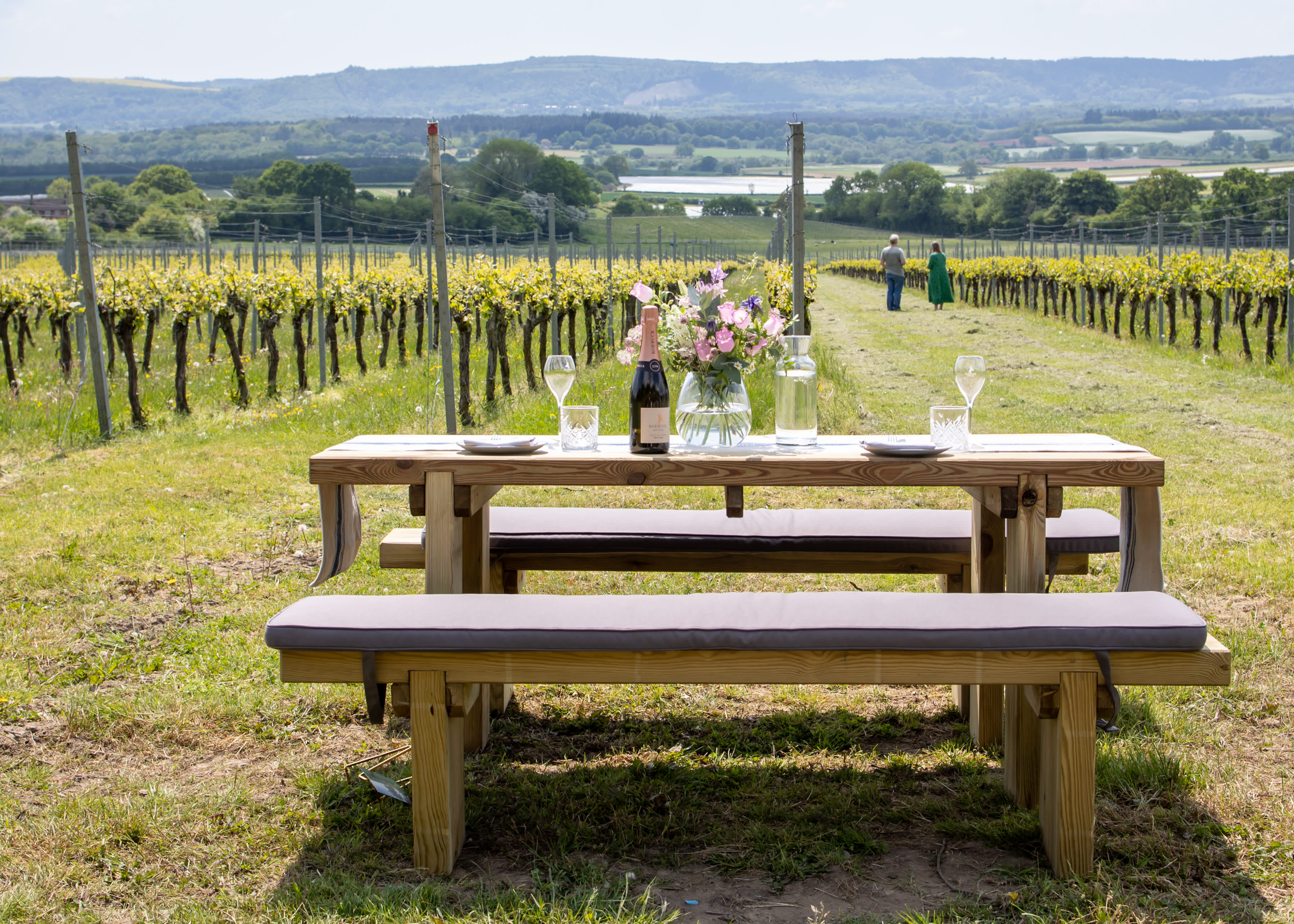 A bottle of English sparkling wine and flowers on a picnic bench at Roebuck Estate vineyard, with vines in the distance