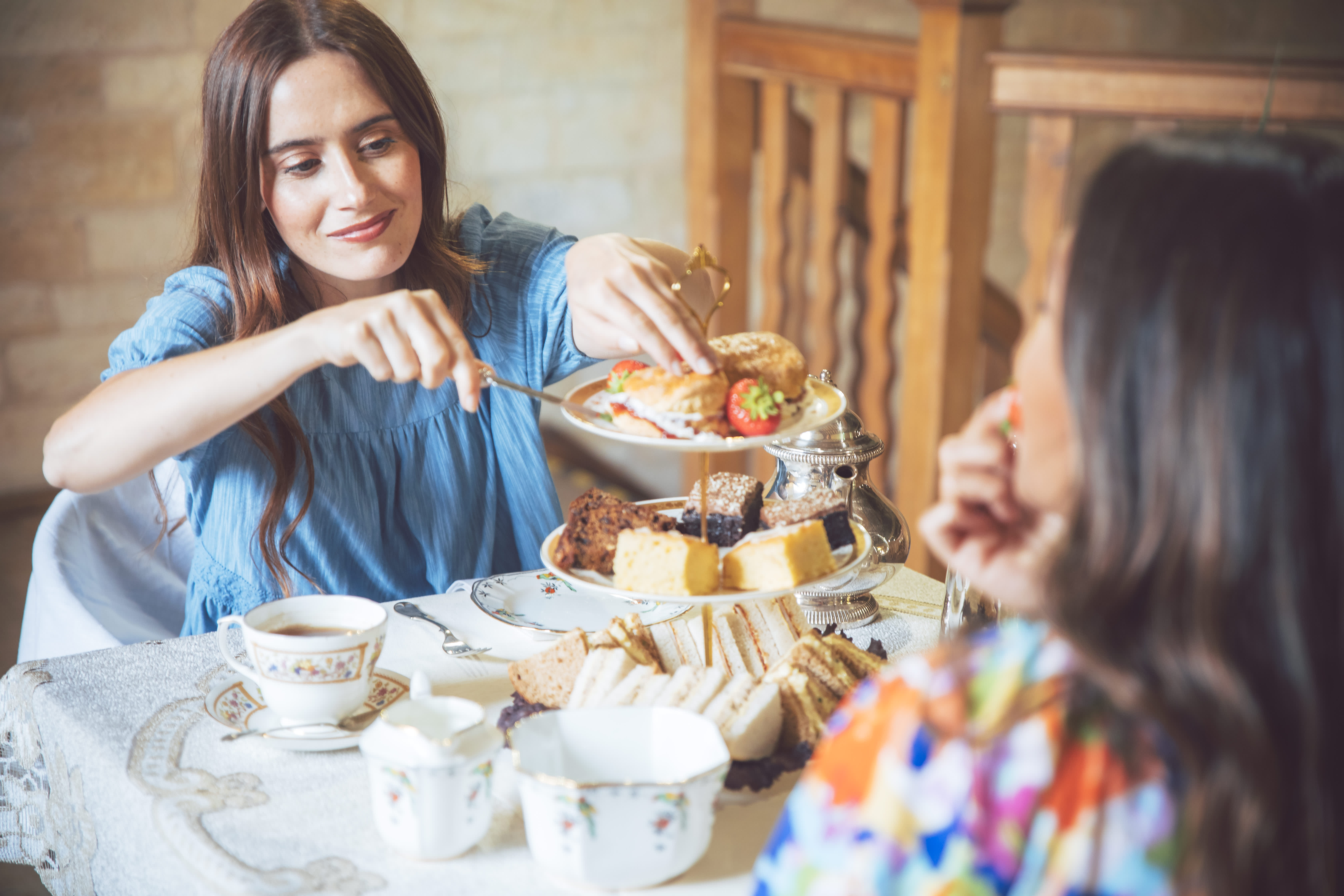 A woman in a blue dress takes a scone from a cake stand, which is filled with sandwiches, scones and cakes, in the Cotswolds