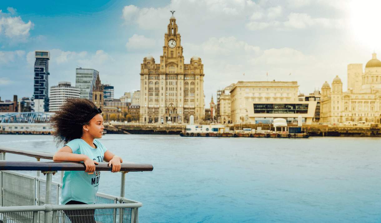 A young child on the Mersey Ferry. They are standing on the end next to the railings looking out over the river. In the background is the Three Graces, the royal Liver Building, Cunard Building and the Port Of Liverpool building.