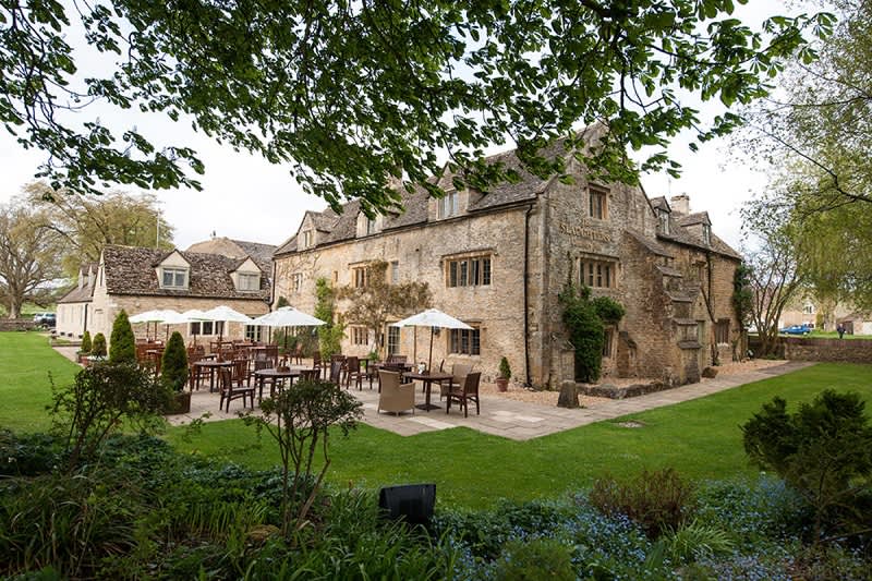 Wooden tables and chairs with white parasols sit on a patio behind The Slaughters Country Inn, surrounded by gardens