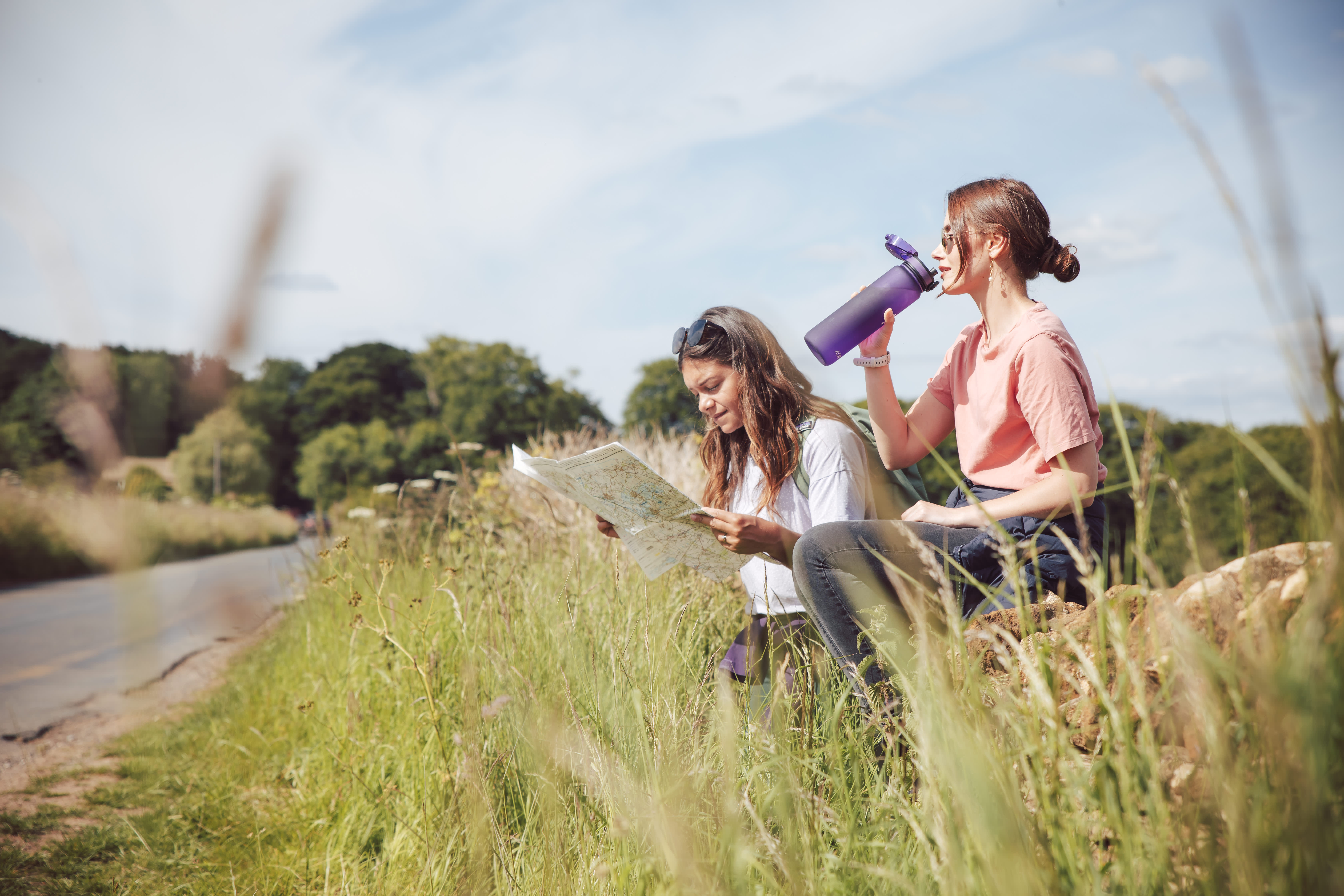 Two women taking a break from walking, one sitting on stone wall drinking from a water bottle, the other reading a map