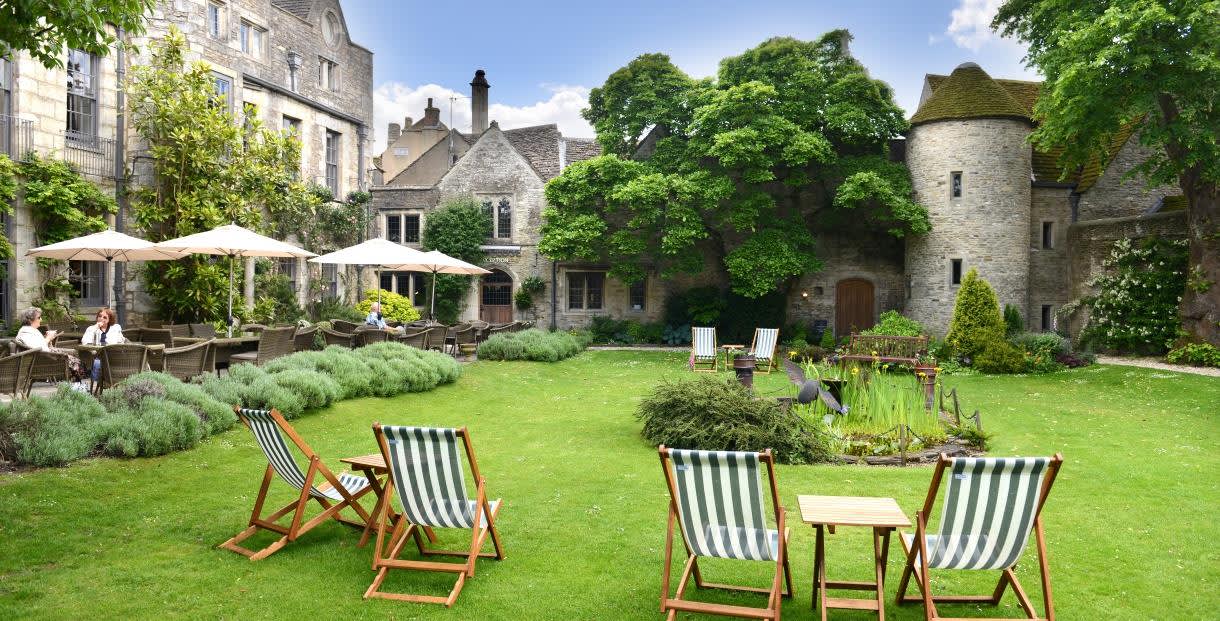 Traditional deckchairs sit on a lawn with flower borders. People sit drinking under sunshades on a patio in front of the 16th century town house at The Close Hotel in the Cotswolds