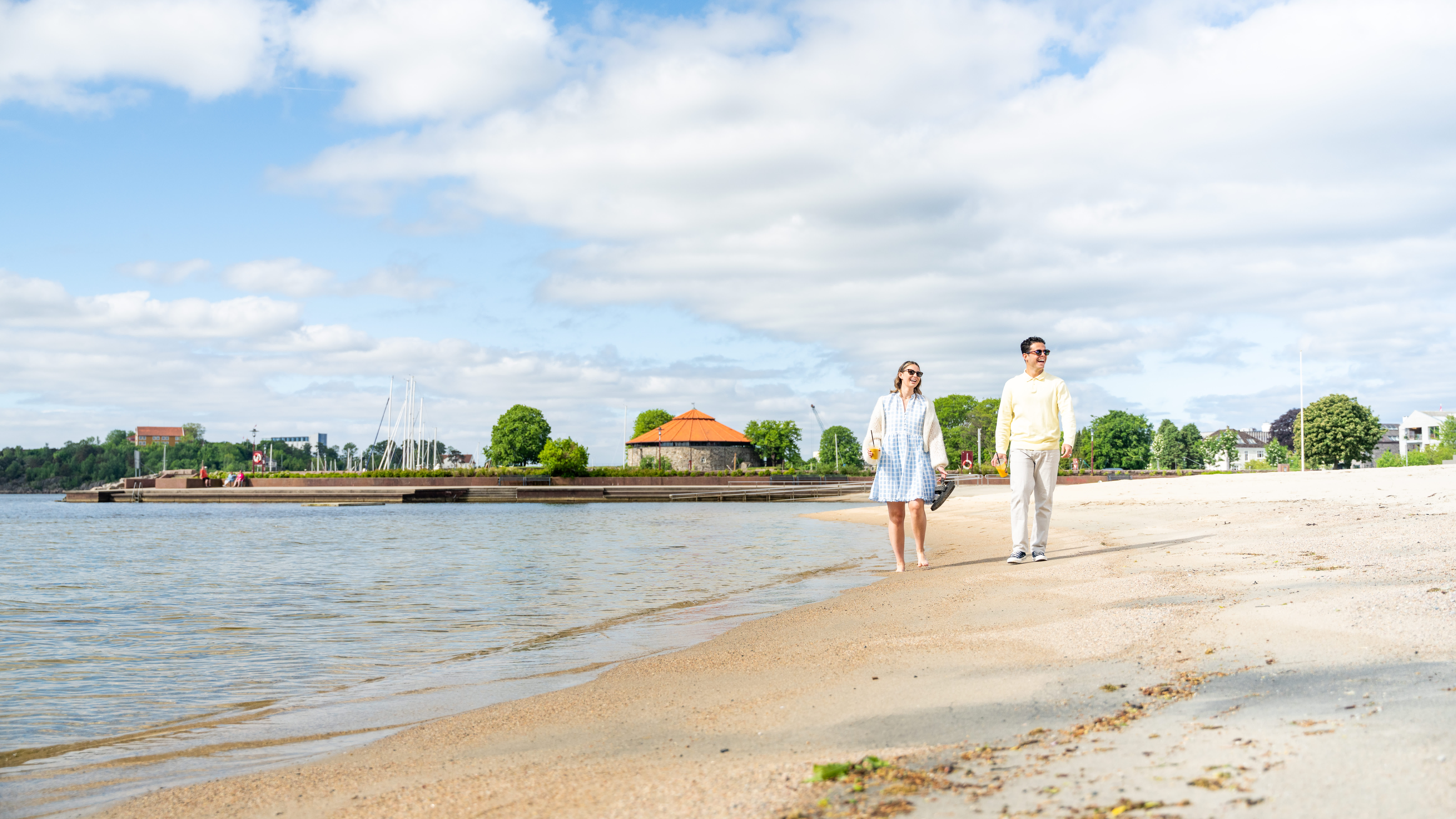 En mann og en kvinne går på tur langs vannkanten på Bystranda i Kristiansand i solskinn