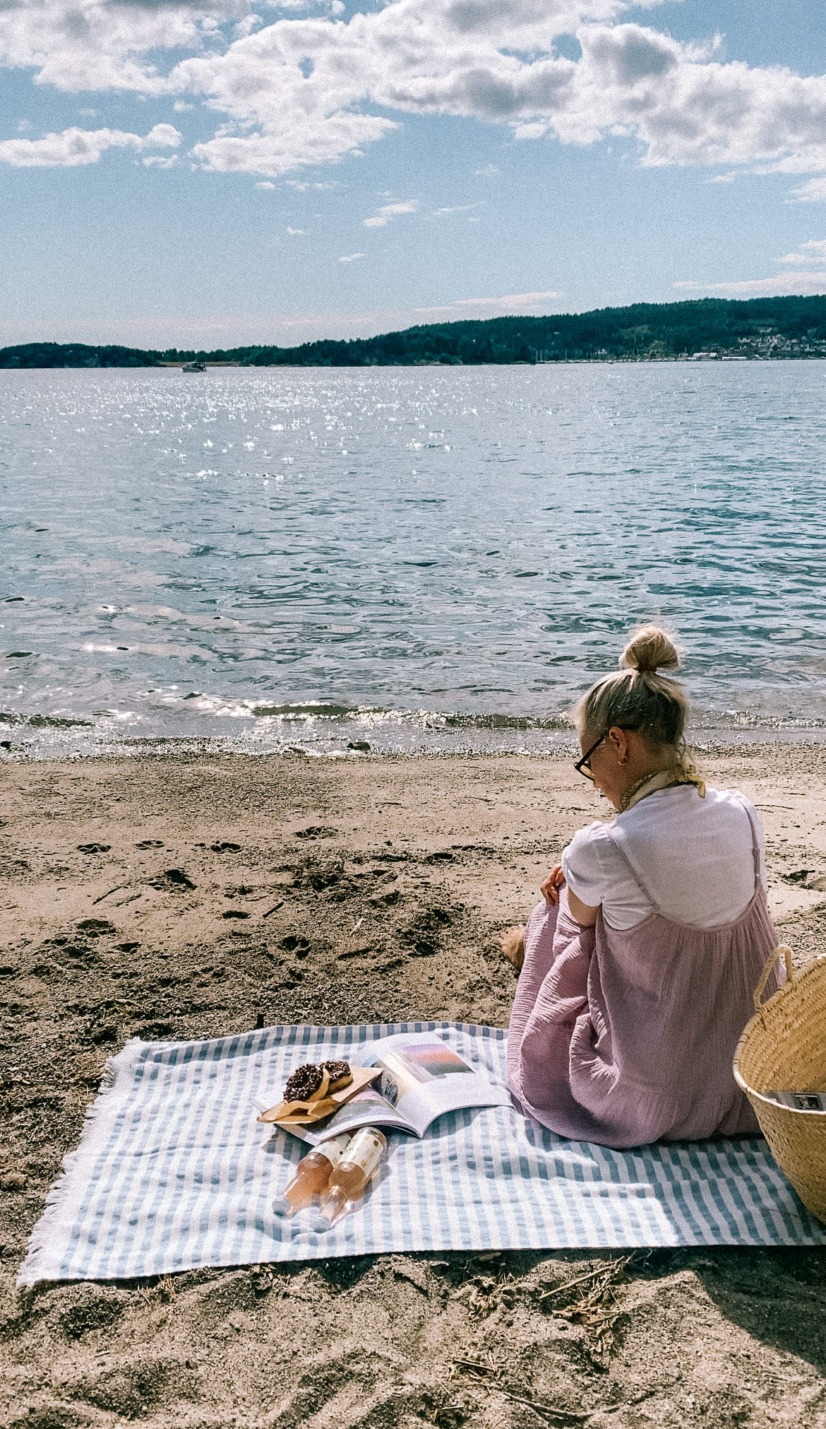 Kvinne som sitter på stranden på et badehåndkle med mat og drikke. Foto.