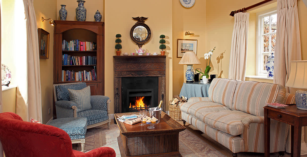 An eclectic mix of sofas and chairs surround a blazing fire with wooden surround in a cozy snug at Bruern Cottages in the Cotswolds