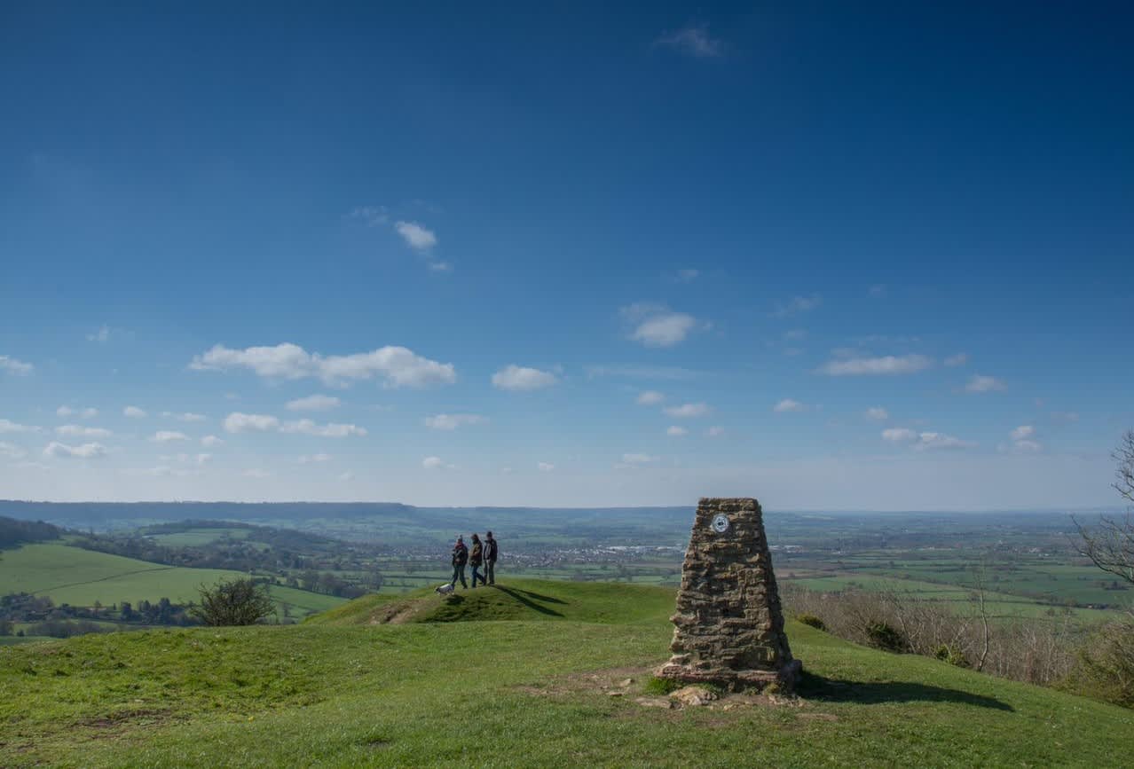 A group of people are standing looking out across rolling green fields under a blue sky with wispy clouds