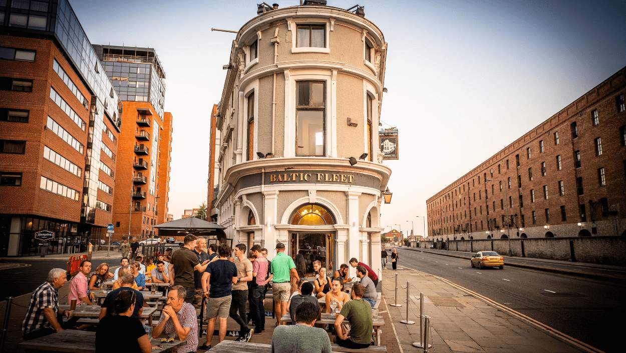 A summer evening outside the Baltic Fleet Pub. The narrow stand-alone pub building is centre of the shot, with the dock warehouse buildings to the right and modern apartments to the left. People are dressed in summer clothes sitting at wooden picnic benches.  i