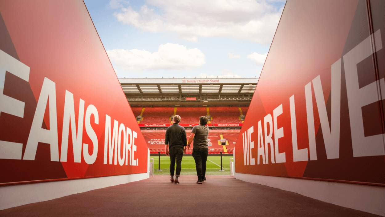 Two people are walking out of the players tunnel at Liverpool's stadium, Anfield. Either side of them are walls in red which have the words 'We are Liverpool. This means more'. In front of the two figures you can see the stand on the other side of the pitch and blue sky with few clouds.