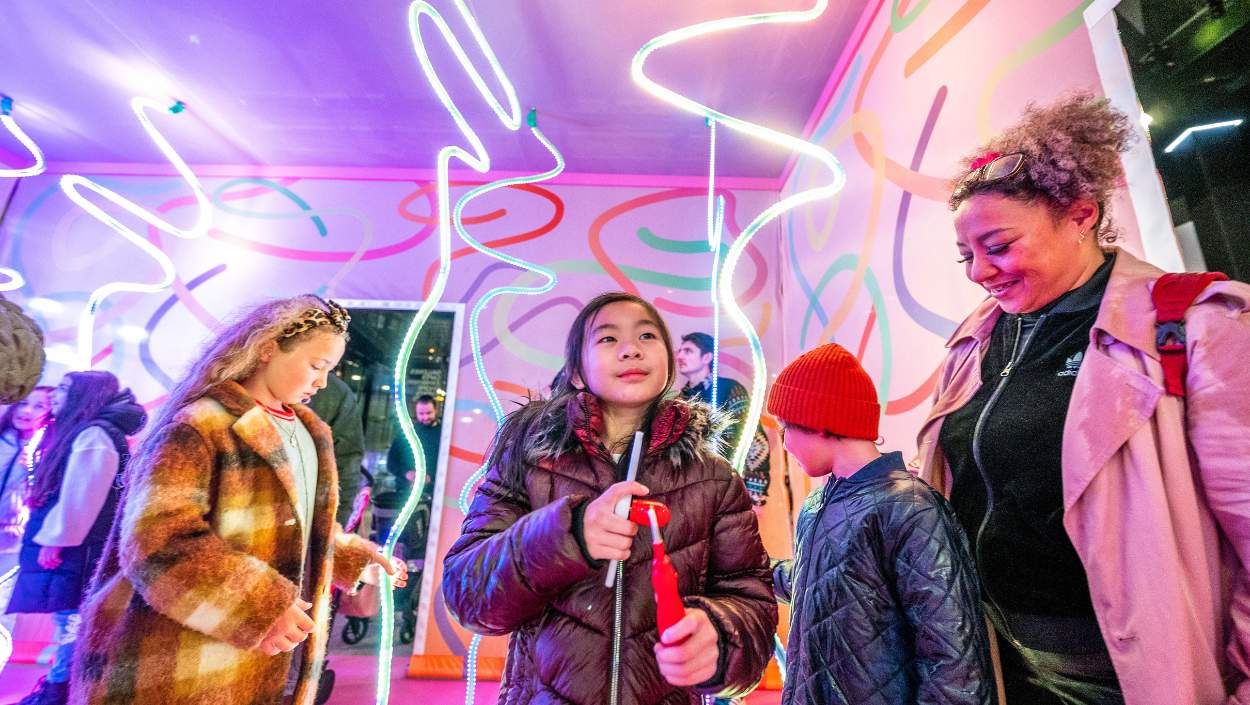 Inside a colourful pink box. There are ribbon shaped LED lights hanging from the ceiling. Children are smiling looking at the lights and an adult is watching the children smiling.