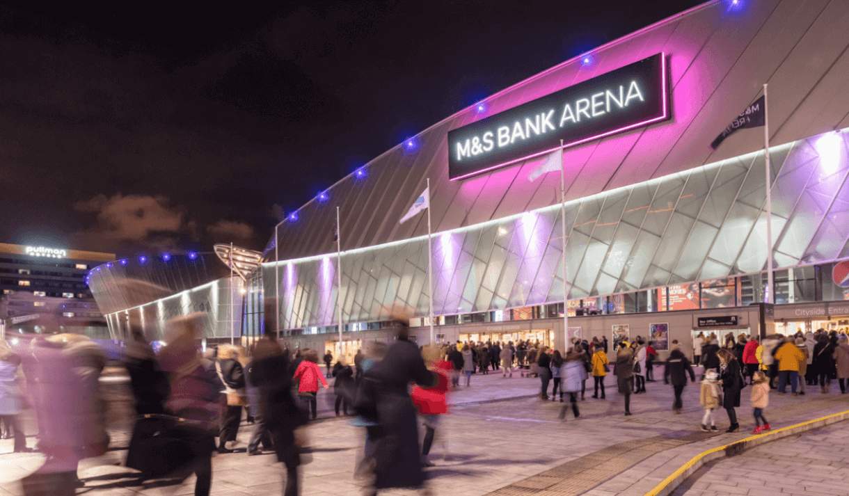 Outside of the M&S Bank Arena with crowds of people walking towards it. The M&S Bank Arena sign with lit up purple.