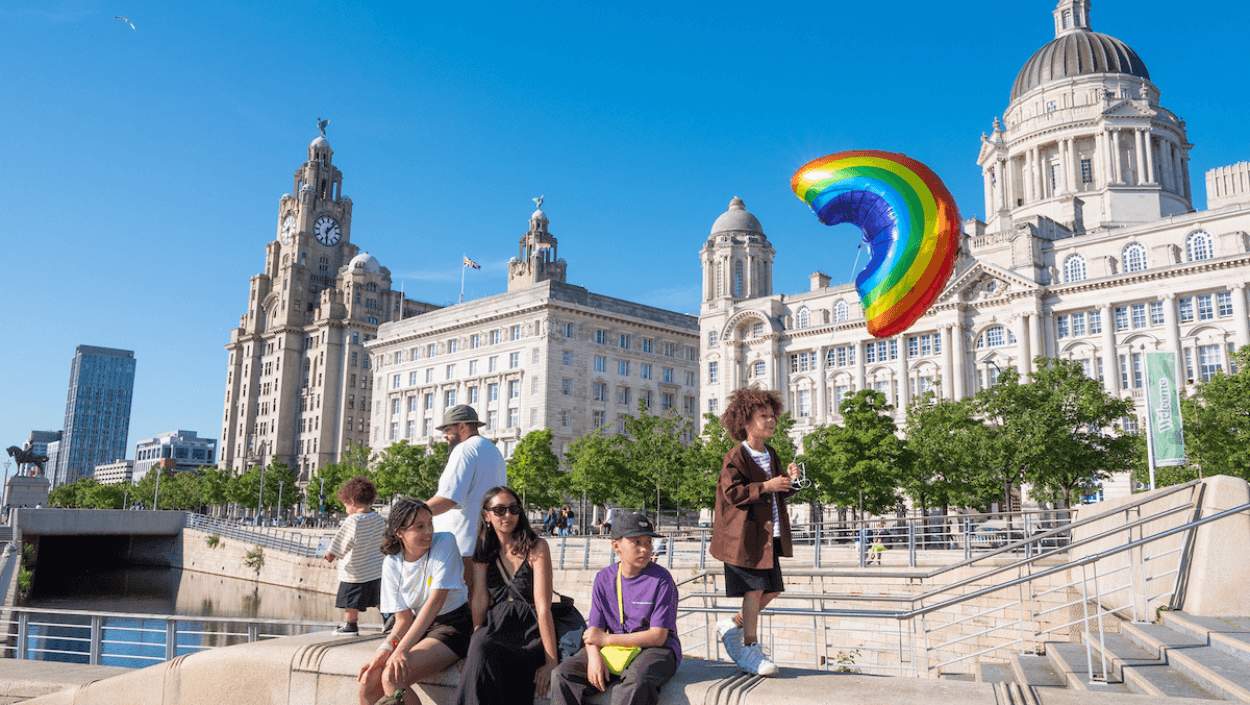 A family on the Liverpool Pier Head on a sunny day. A boy holding a rainbow balloon in front of the three graces buildings.