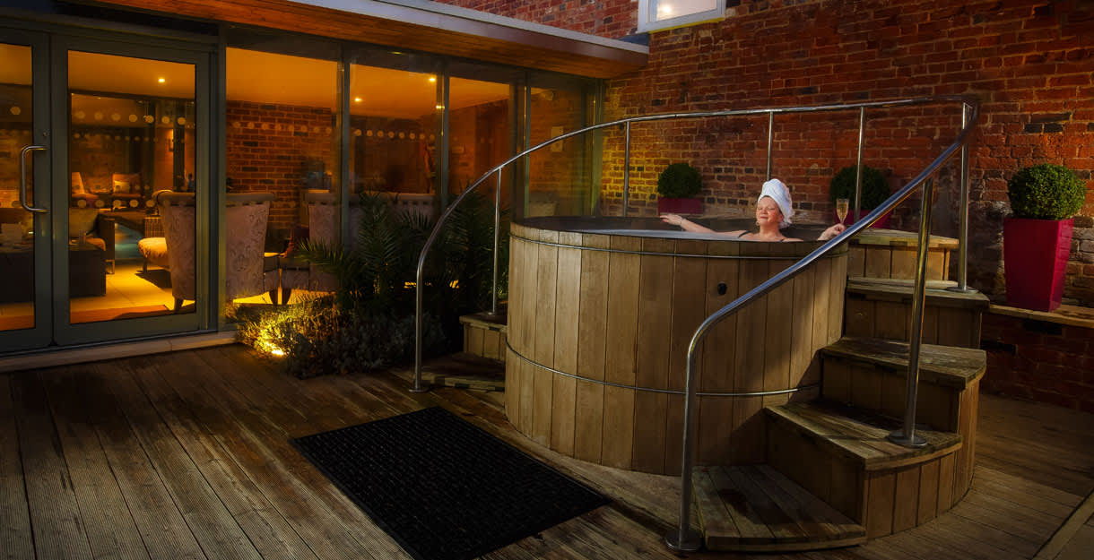 A lady relaxes at night in an outdoor wooden hot tub, surrounded by plants and subdued lighting at The Greenway Hotel & Spa in the Cotswolds