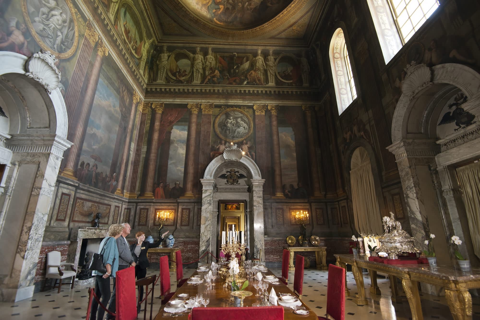 A couple with a tour guide stand in an impressive room with painted images on the ceiling and walls, large marble doorways and gold furniture at Blenheim Palace
