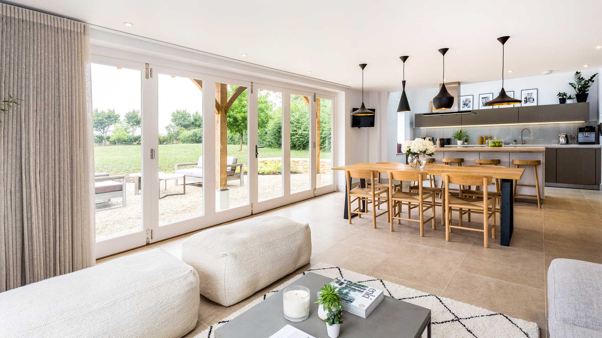 An open plan living space with modern kitchen, wooden dining table and elegant seating area, opening out through bi-fold doors to a patio. StayCotswold in the Cotswolds