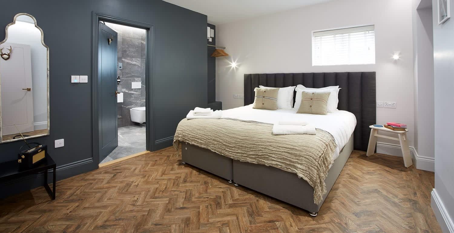 Hotel room with a double bed in the centre with cream and white bed linen. The floor is wooden parquet. One wall is dark grey with a door to the bathroom. The other two walls are white,