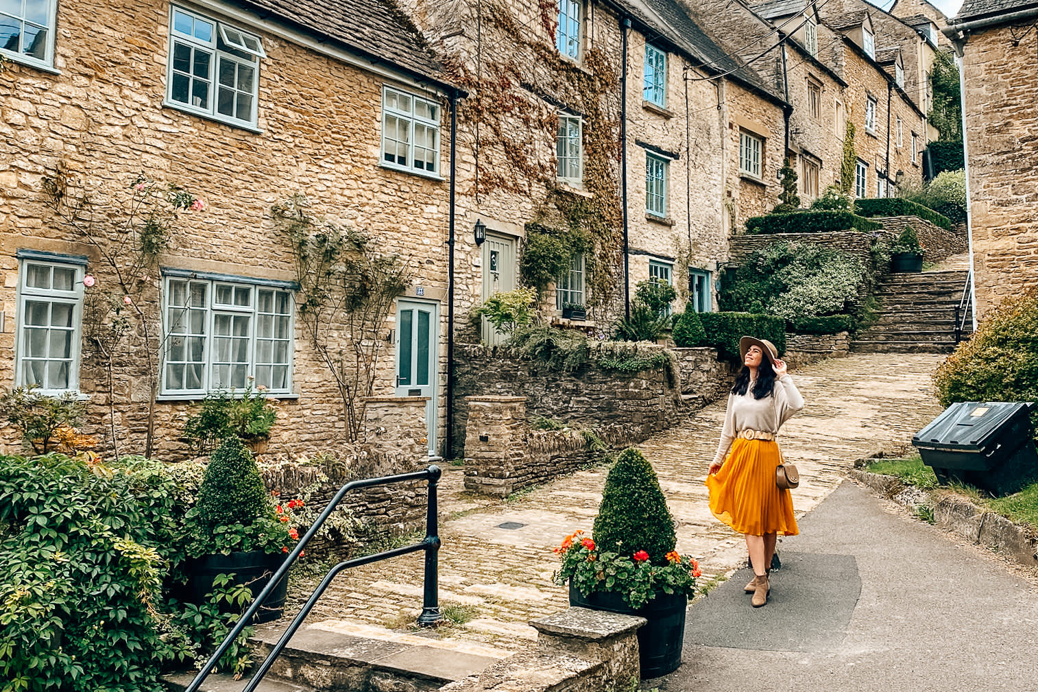 A woman wearing a hat smiles as she poses in front of stone cottages on a stepped hill in Tetbury