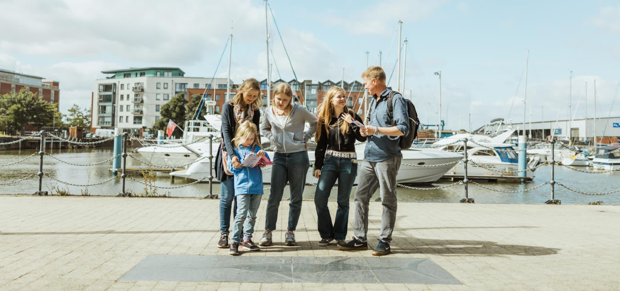 A group of people standing beside a stone fish carving on Hull’s Fish Trail near the marina, holding trail maps with boats and water in the background.