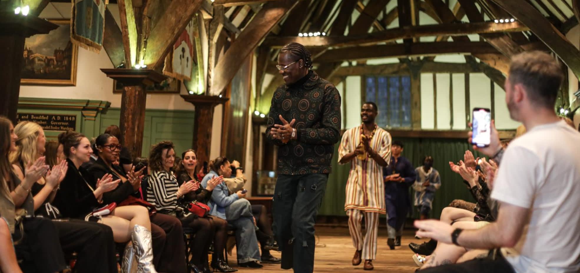 Seguntuyi Onakoya walks down a runway at Ferens Art Gallery whilst the audience claps. He smiles at the onlookers and a male model follows him on the runway.