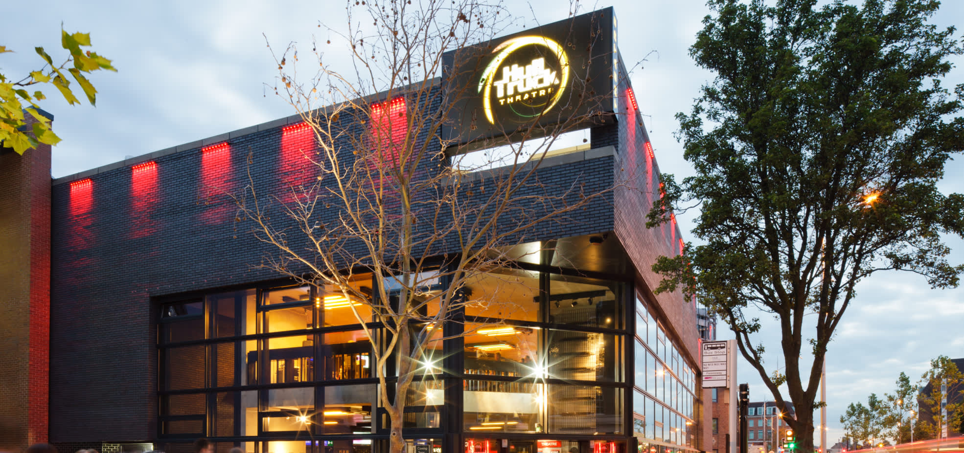 Exterior view of Hull Truck Theatre at dusk, showing the modern brick building illuminated with red lighting and large glass windows, with the theatre’s bright circular logo displayed above the entrance and trees framing the scene.