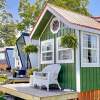 Green vertical-sided tiny house with a small front porch furnished with white wicker seating, set against a blue sky and leafy green tree.
