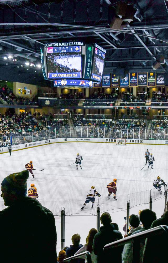 Fans watch a college hockey game between the University of Notre Dame and Minnesota at Compton Family Ice Arena. The ice rink is surrounded by a full crowd, with players skating mid-game and a large scoreboard hanging above the ice.