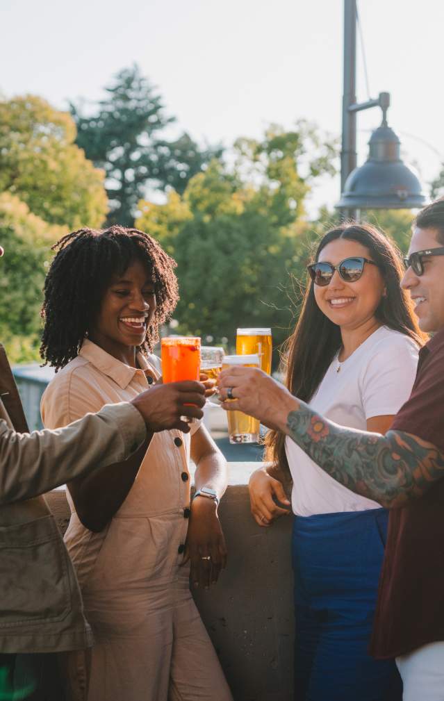Four adults perched on a rooftop patio cheers their beverages with smiles and a happy atmosphere.