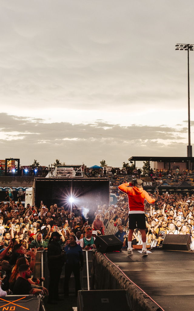 A performer in a red hoodie stands on an outdoor stage facing a massive crowd at sunset. Thousands of fans pack the venue, many holding up phones to capture the moment, while bright stadium lights and food stand signs glow in the background.