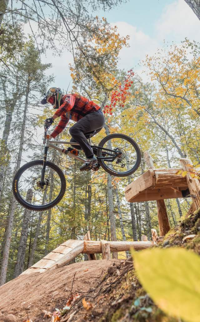 Mountain biker in red gear launches off a wooden ramp, airborne among tall autumn trees, conveying adventure and exhilaration.