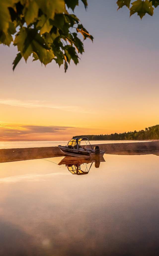 A serene sunset over a calm Lake Superior, framed by tree leaves. A small boat rests on the shore, with its reflection mirrored in the still water.