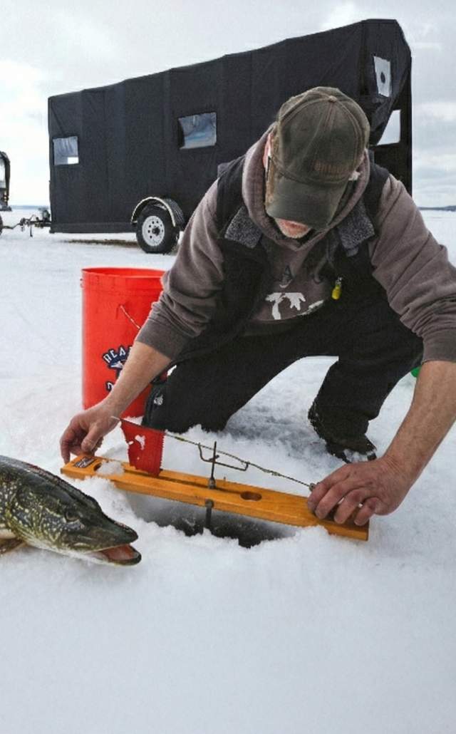 Man ice fishing on a frozen lake in the Upper Peninsula of Michigan with a large Northern Pike lying on the snow beside the hole.