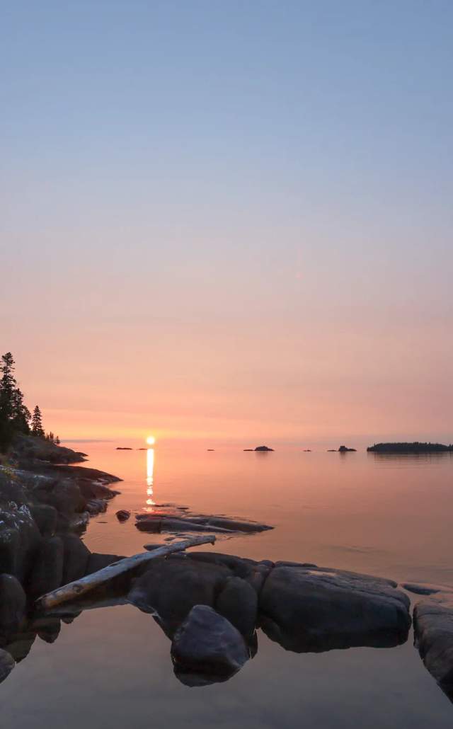 A person in a white jacket sits on rocks by a calm lake, gazing at a sunset with pastel colors reflecting on the water and silhouetted trees in the background.