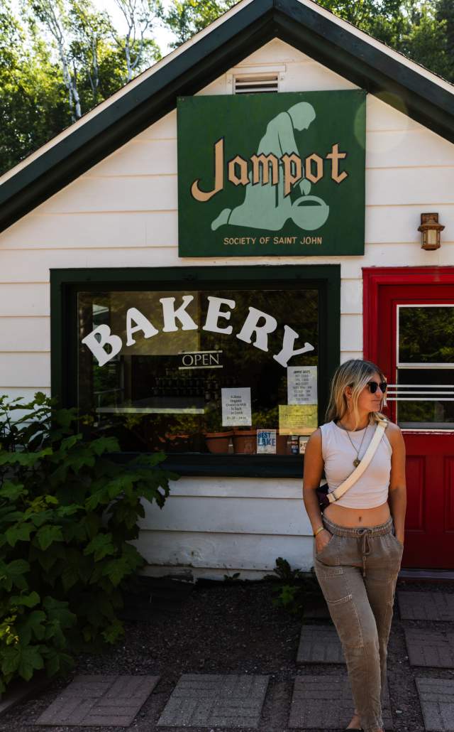 A young woman in a white crop top and beige pants stands in front of a small bakery named "Jampot," which has a green sign and a red door. The window displays large letters spelling "BAKERY" and jars of baked goods inside.