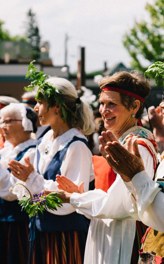 A group of people in traditional clothing, wearing floral wreaths, clap and smile at an outdoor event under sunny skies, conveying joy and celebration.