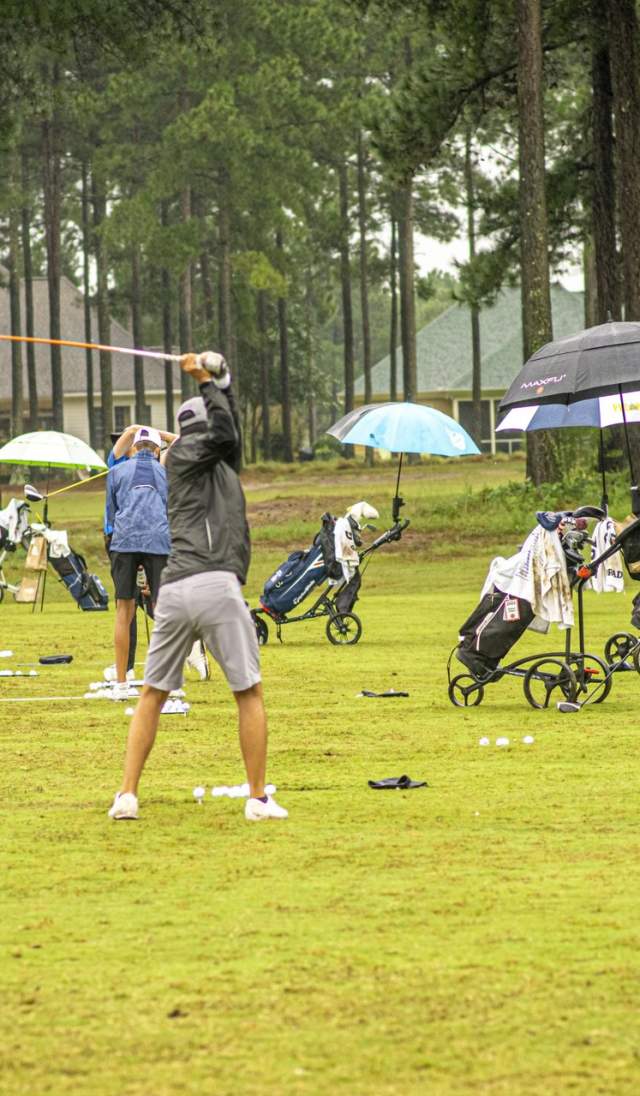 Golfers warm up on a managed practice area with caddies, equipment carts, and coordinated spacing, reflecting a tournament-ready course environment.