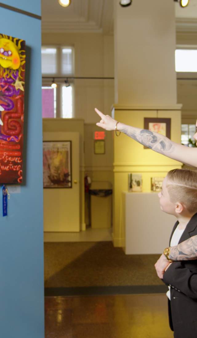 Adult and child viewing a colorful art exhibit inside a local arts center, with the adult pointing at the artwork on display.
