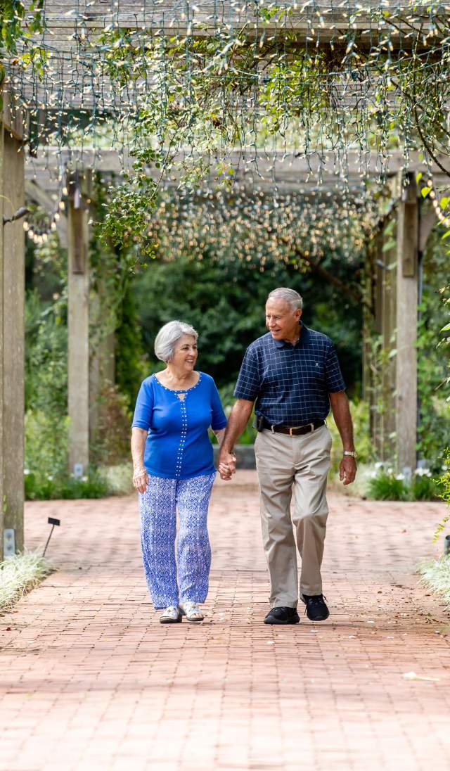Couple walking together along a brick pathway beneath a vine-covered pergola in a botanical garden