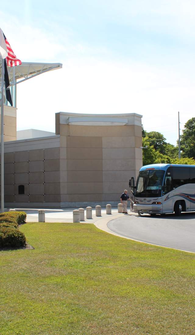 Motorcoach bus parked outside the U.S. Army Airborne & Special Operations Museum in Fayetteville, North Carolina, with flags and entrance visible on a sunny day