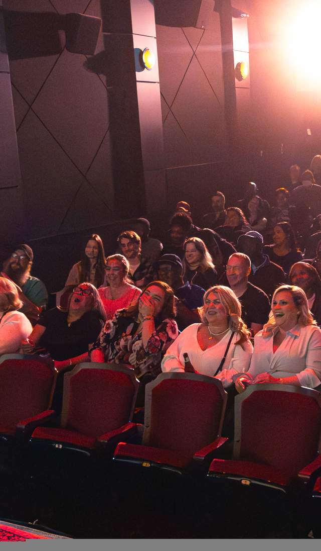 Stand-up performer on stage facing a packed theater audience, who are laughing under bright stage lights.