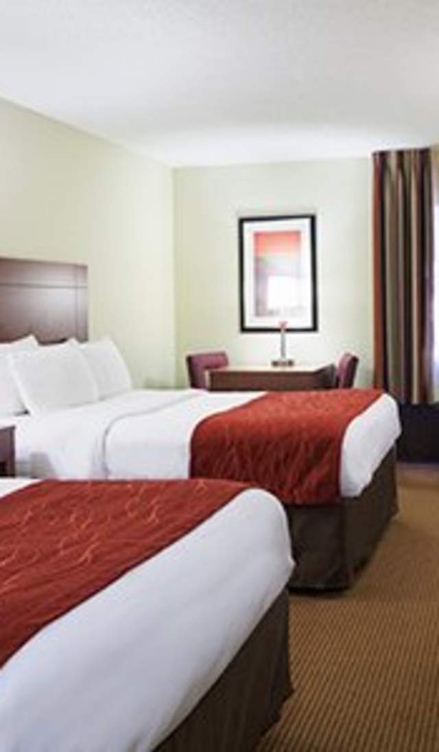 Hotel room with two beds featuring white bedding and red accent throws, wood headboards, bedside tables with lamps, a desk, dresser with TV, and a window with curtains and an in-room air-conditioning unit.