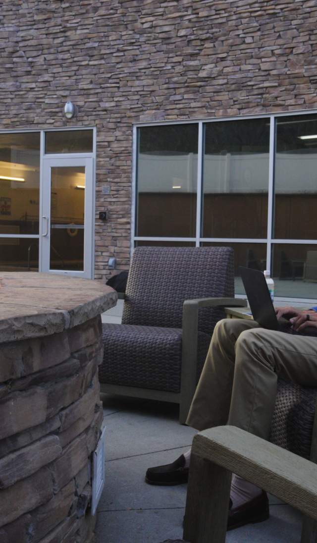 Person sitting in an outdoor courtyard lounge chair beside a stone fire pit with visible flames, surrounded by modern seating and large windows on a brick building.