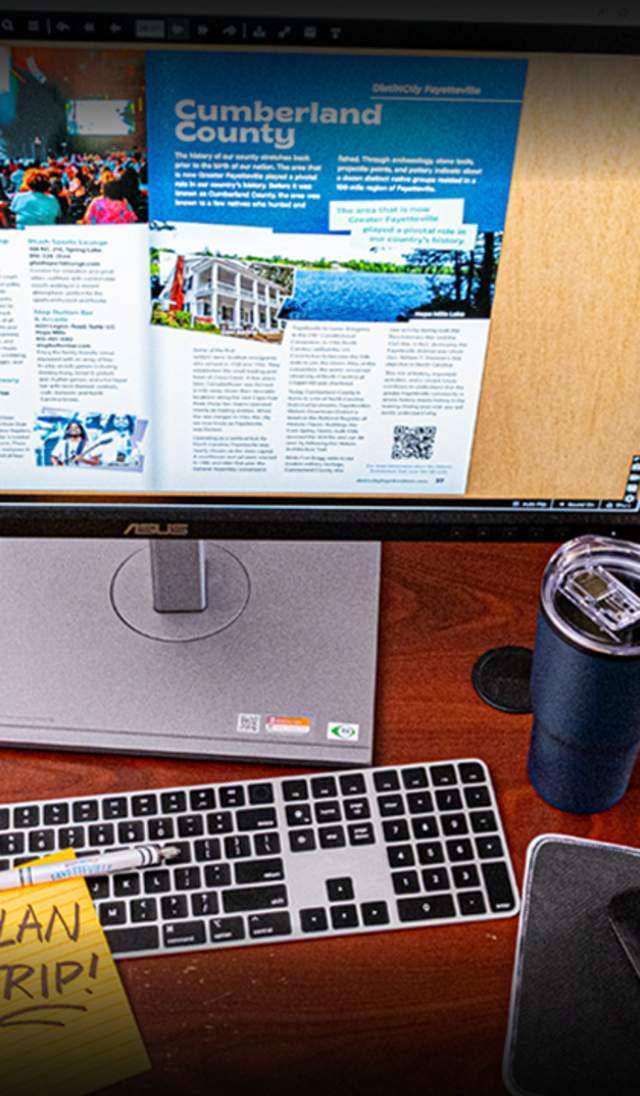 A digital destination guide is open on a computer monitor at an office desk with a keyboard, mousepad, a blue travel cup, and a yellow sticky note that says Plan Trip.