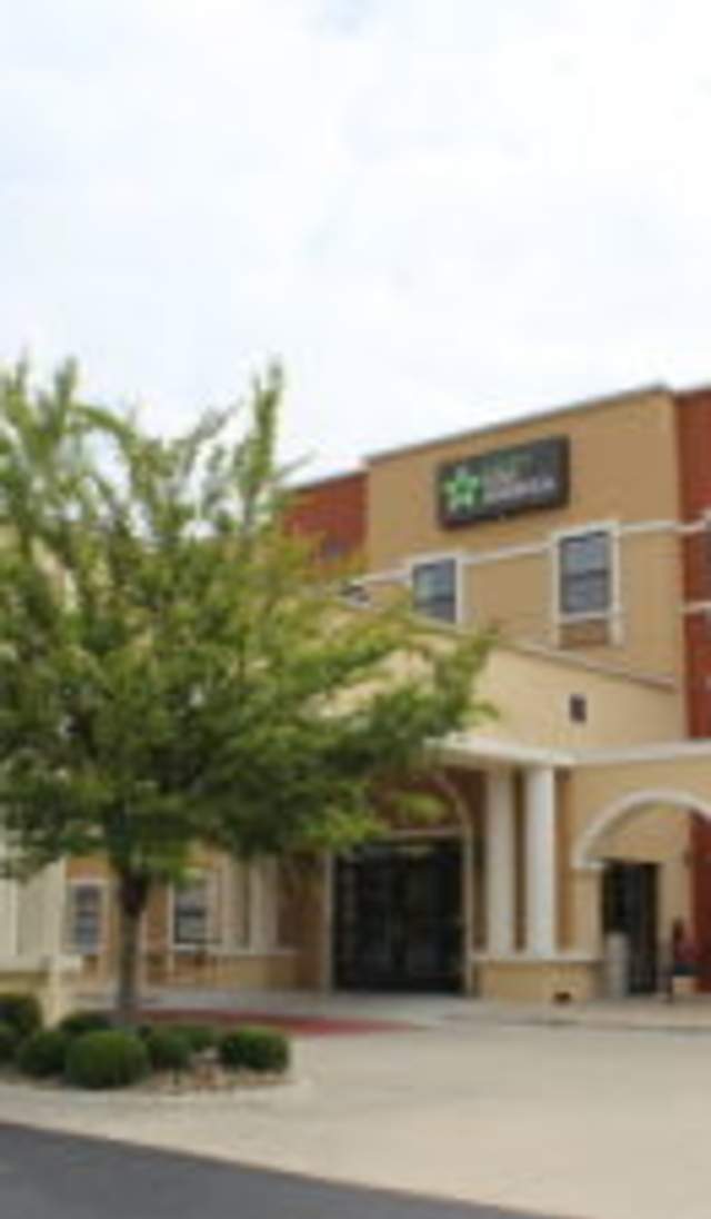 Exterior view of a multi-story hotel with arched entrances, tan and brick-colored facade, landscaped trees and shrubs, and a covered drop-off area at the main entrance.